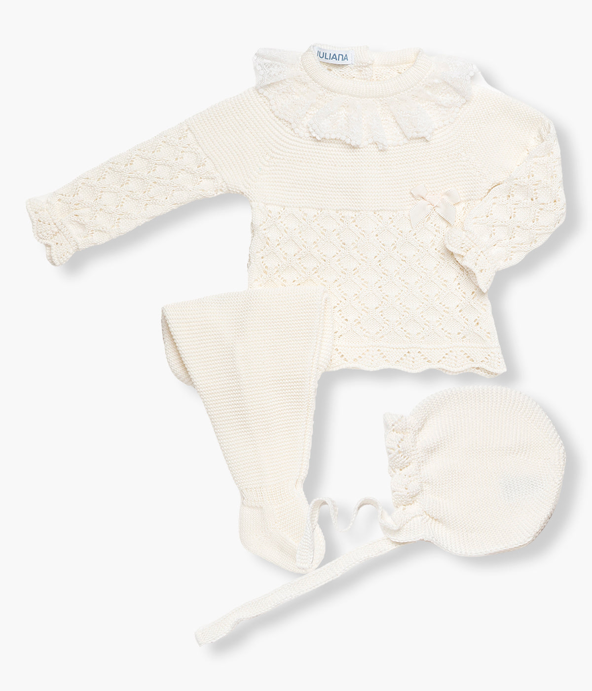 Vista de frente do conjunto em malha pérola da Juliana para bebé menina e recém‑nascida, composto por camisola, calças e touca. Camisola com gola folho em renda detalhada, detalhes perfurados na malha, rebordo ondulado nos contornos e laço gorgurão decorativo. Touca e calças com pés a combinar.