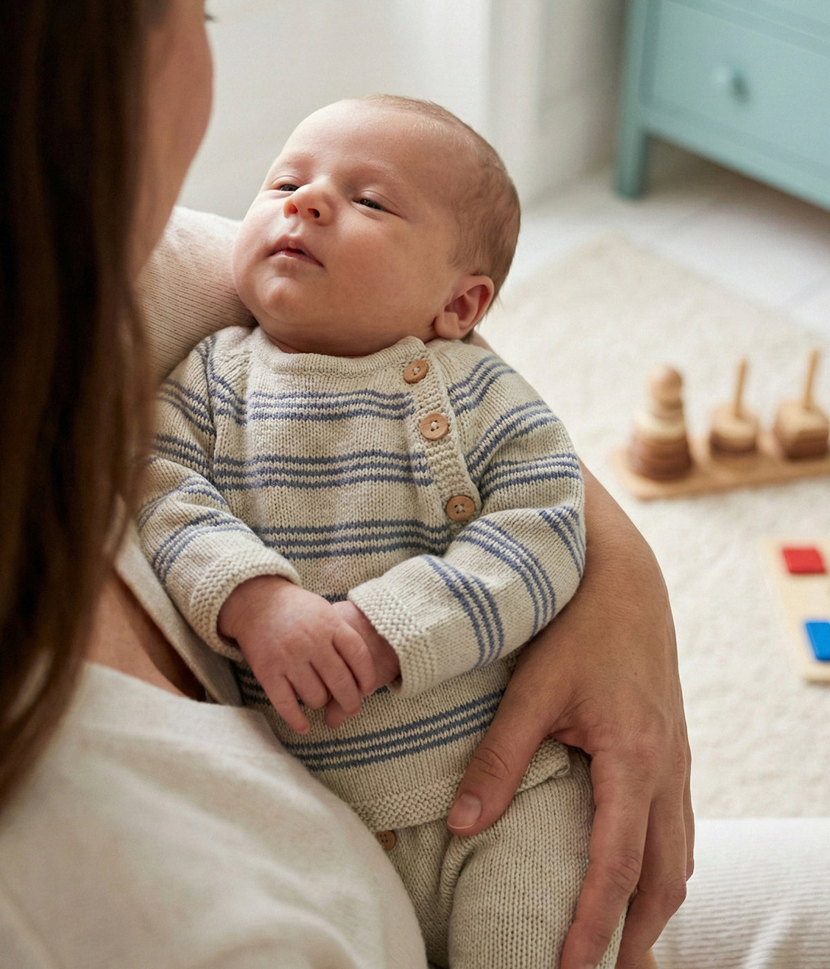 Bebé a vestir o conjunto em malha de algodão orgânico bege da Mini Mooi para bebé e recém‑nascido(a), composto por camisola e calças. Camisola de manga comprida com riscas azuis e abertura no ombro com botões de madeira. Calças lisas com pés com botões de madeira decorativos.