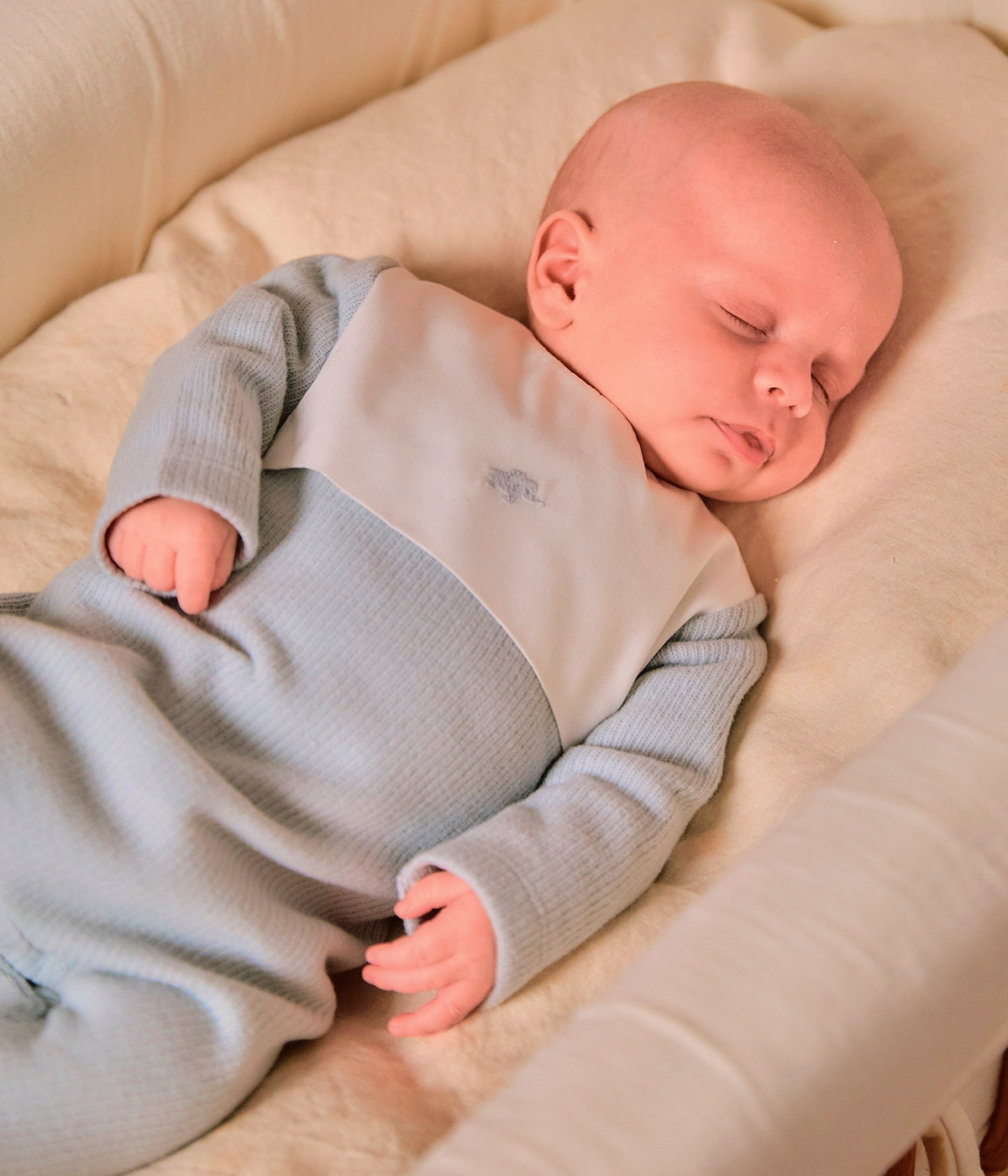 Bebé a usar o babygrow em algodão azul acinzentado da Laranjinha para bebé e recém-nascido(a). Peitilho em tecido branco com elegante bordado de elefante. Abertura nas costas e entrepernas.