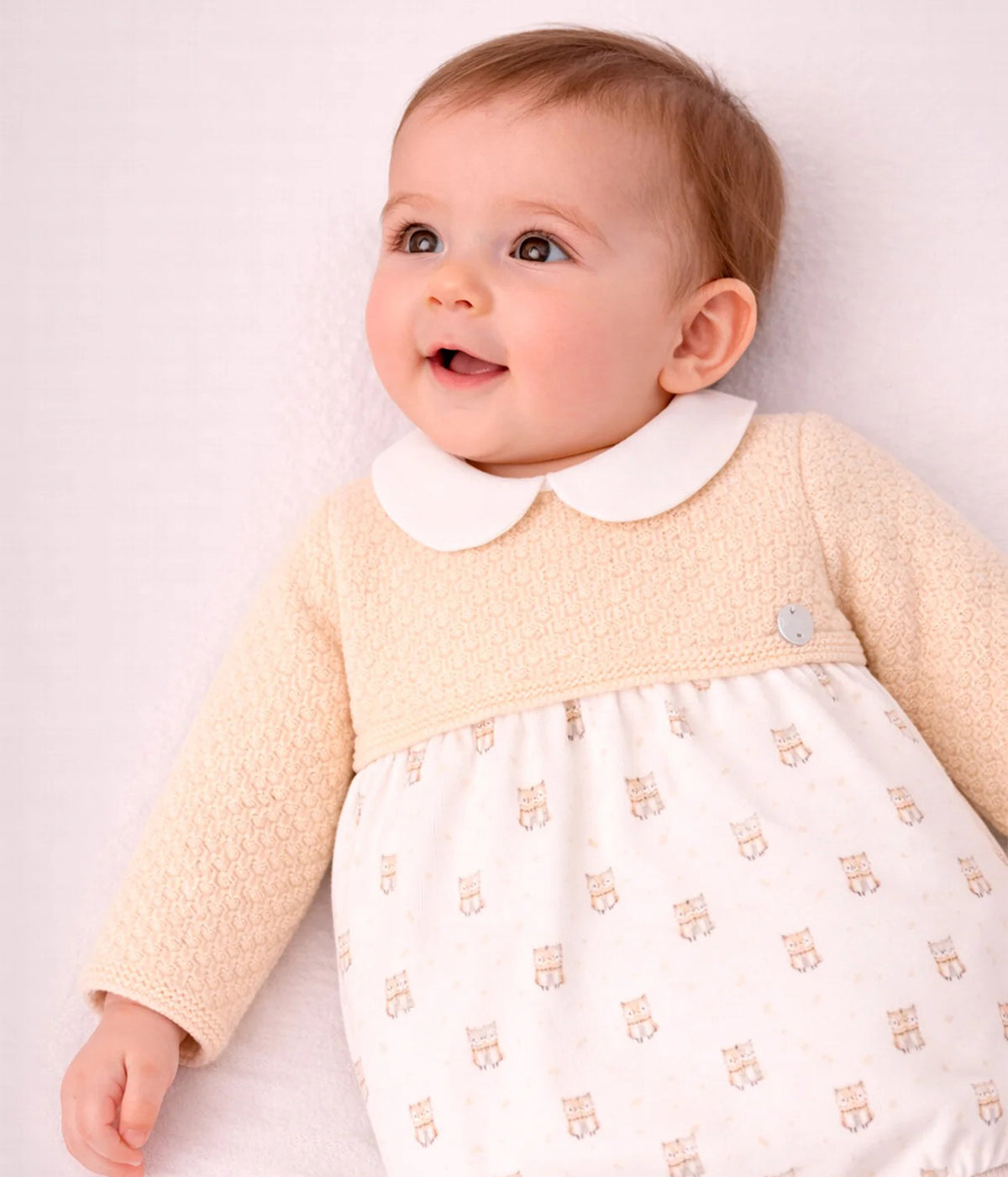 Bebé a vestir o vestido em malha e algodão da Paz Rodríguez para bebé menina e recém-nascida. Parte superior em malha detalhada em tons de amarelo com gola folho branca com picueta bege. Parte inferior em tecido de algodão branco com padrão gatos. Tapa fraldas a combinar. Abertura nas costas com botões.