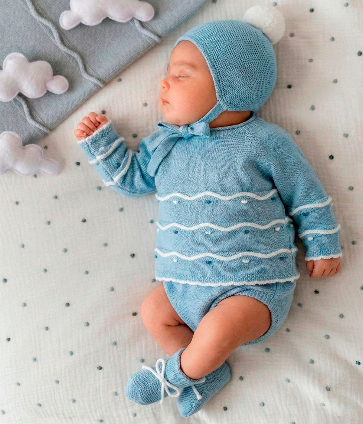 Vista com o bebé do conjunto de 4pc em malha da Mac Ilusión para bebé e recém-nascido(a). Camisola com detalhes com relevo em branco e tons de azul. Tapa fraldas liso. Gorro com pompom branco. Carapins tipo bota.