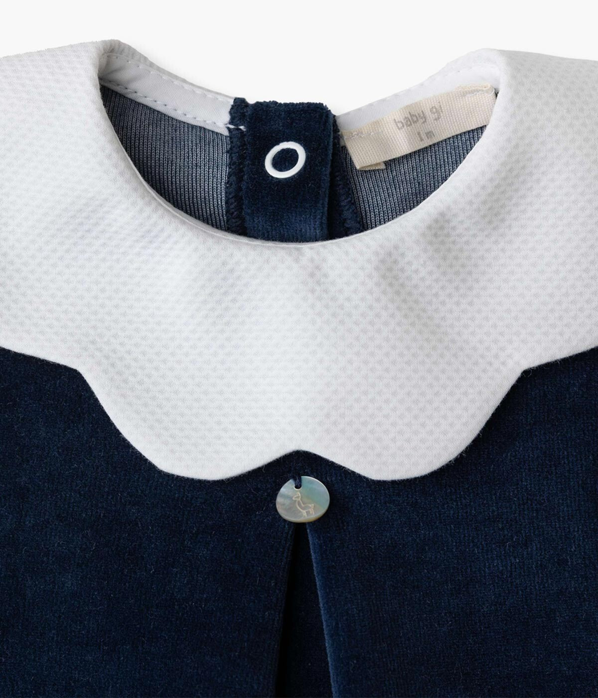 Babygrow em veludo azul-marinho da Baby Gi para bebé e recém-nascido(a). Gola ondulada branca e vinco com medalha Baby Gi na frente. Abertura com molas de pressão nas costas e entrepernas.