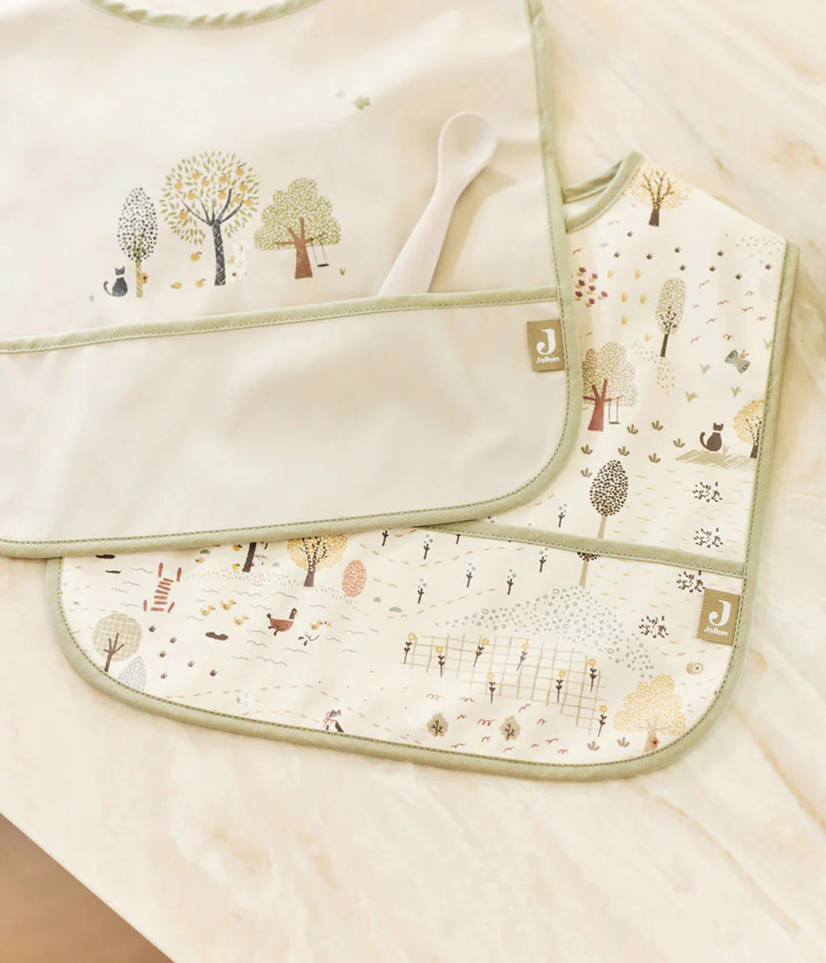 Conjunto de duas babetes impermeáveis com padrão árvores e animais coleção Tiny Park da Jollein para bebé e recém-nascido(a). Babetes impermeáveis com bolso, uma lisa verde e outra pérola, perfeitas para manter o bebé limpo enquanto aprende a beber e comer sozinho.