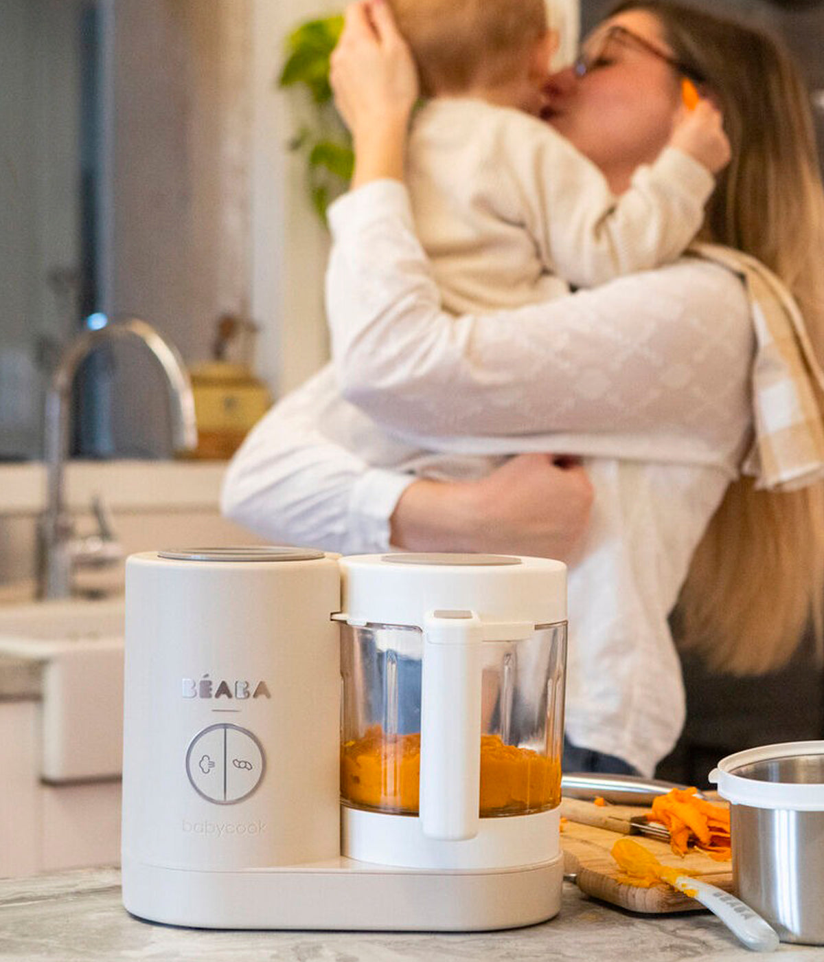 Vista com bebé do robô de Cozinha Babycook Néo da Béaba na cor Clay Earth (bege). Inteligente robô de cozinha premium para bebé 6 em 1 que cozinha a vapor, tritura, descongela, aquece refeições, aquece biberões e esteriliza acessórios.