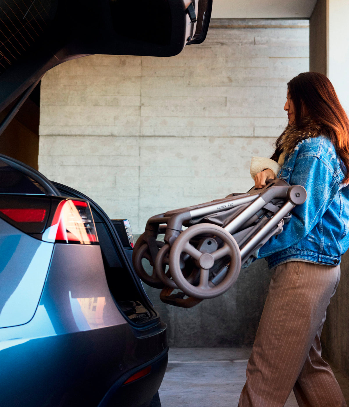 Mulher a colocar o carrinho de passeio Flo da marca Anex na cor Yuki na bagageira de um Tesla.