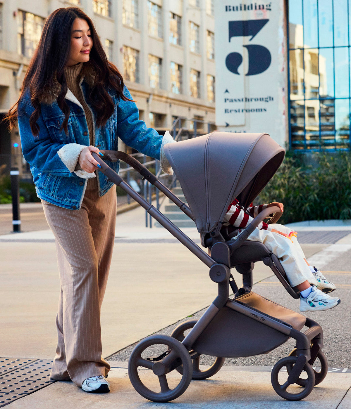 Mulher a utilizar o carrinho de passeio Flo da marca Anex na cor Yuki. Elegante carrinho que acompanha o bebé desde o nascimento até aproximadamente aos 4 anos, combinando uma alcofa leve e espaçosa com um assento de passeio reversível até aos 22 kg.