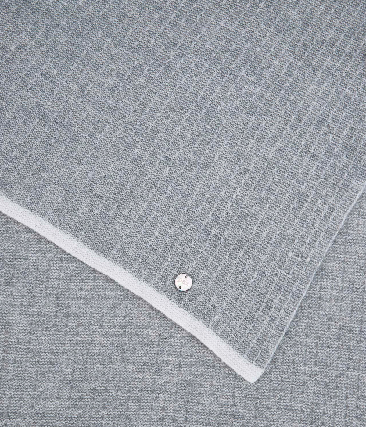Vista detalhada da manta em malha de lã e caxemira cinza da Paz Rodríguez para bebé e recém-nascido(a). Contorno a toda a volta em branco. Medalha da marca em madrepérola.