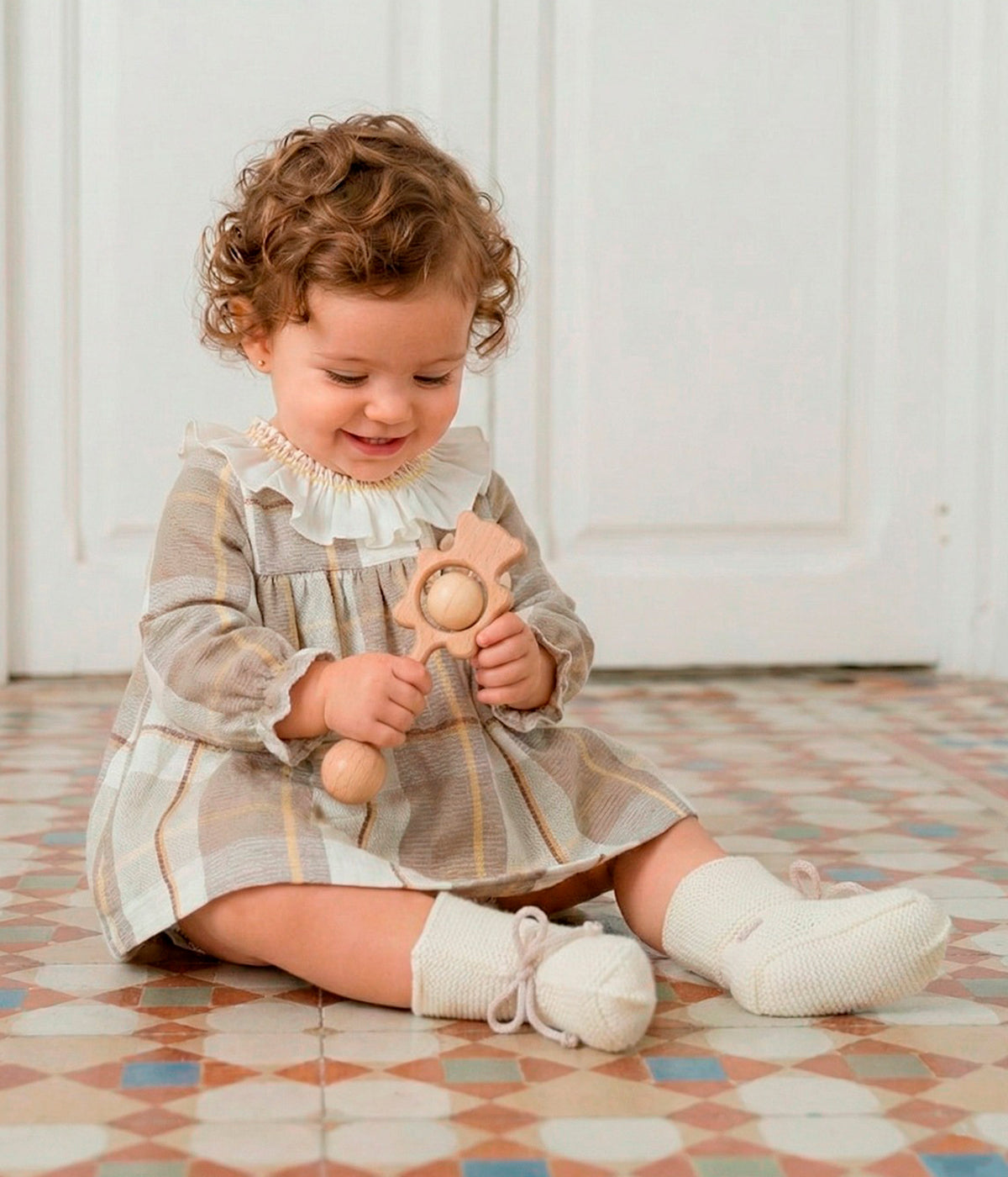 Bebé menina a vestir o vestido em algodão da Mac Ilusión para bebé menina e recém-nascida. Elegante vestido com padrão xadrez em tons de bege e castanho, gola folho branca e pequeno pompom com laço no peito. Tapa fraldas a combinar.