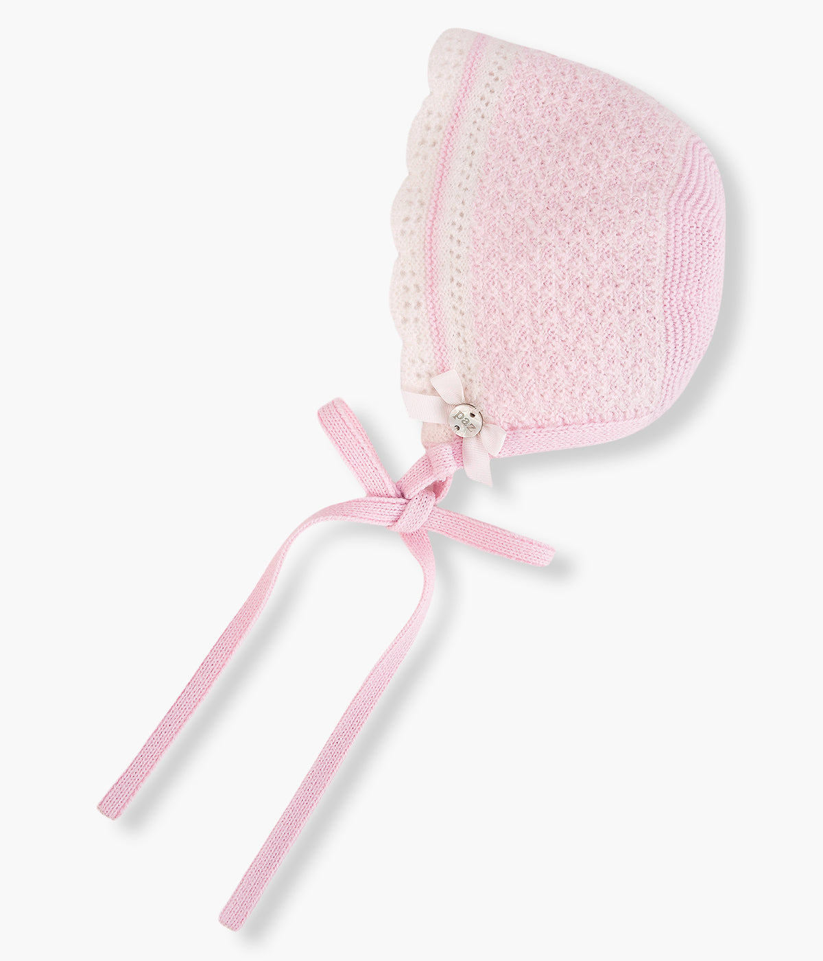 Vista lateral da touca de malha rosa da Paz Rodríguez para bebé menina e recém-nascida. Suave touca com contornos ondulados, detalhes com relevo, medalha de madrepérola com laço e tiras para atar no queixo.
