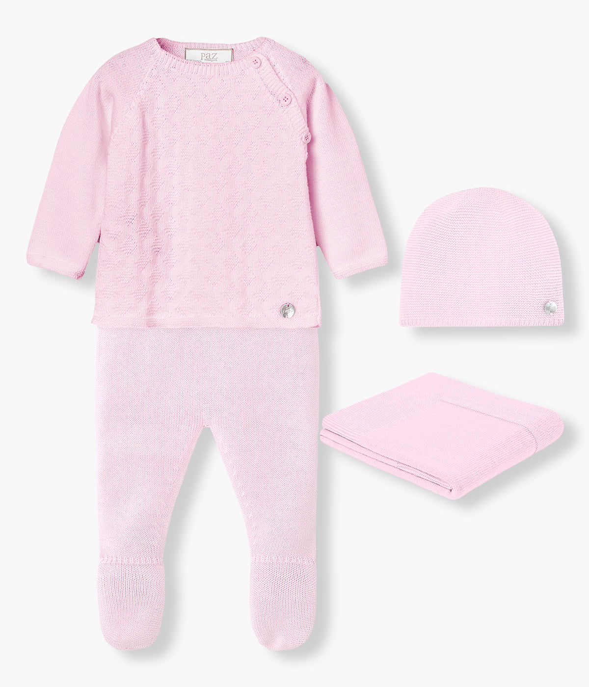 Vista de frente do conjunto de 4 peças em malha de algodão rosa da Paz Rodríguez para bebé menina e recém-nascida. Camisola de abertura no ombro com detalhes na malha. Calças lisas com pés. Gorro liso com medalha madrepérola. Manta simples de formato retangular.